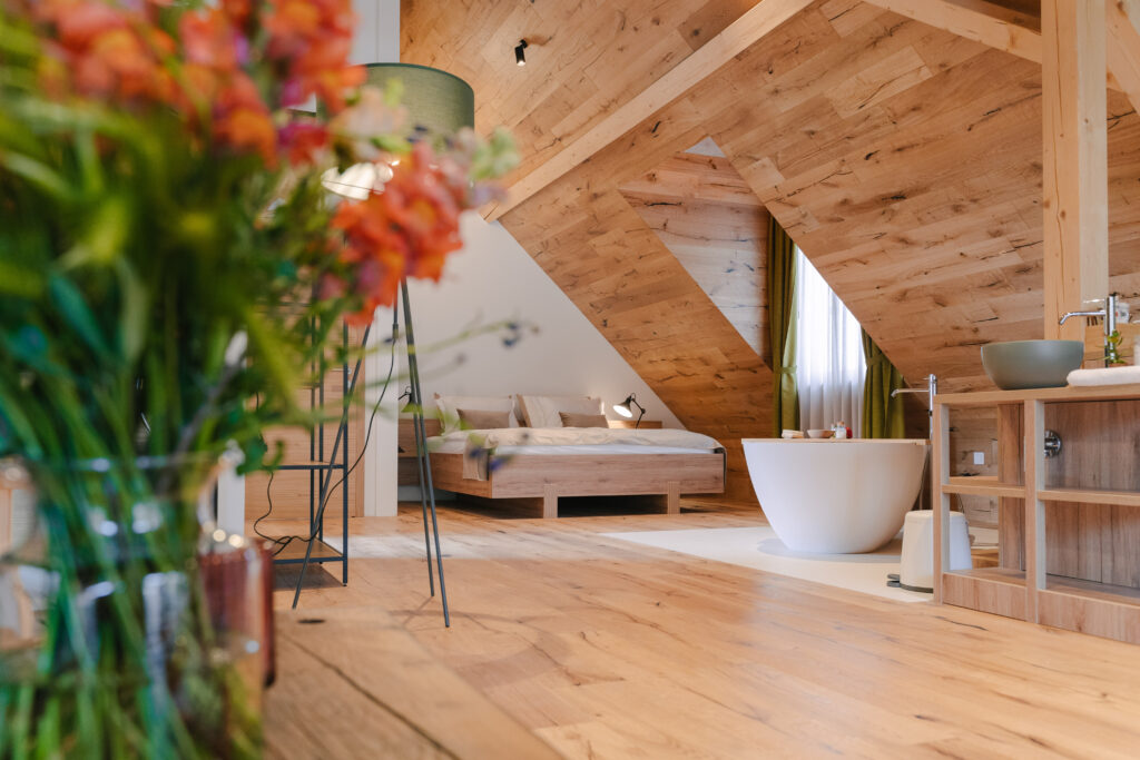 Svetlá podkrovná kúpeľňa a spálňa s detailmi zo svetlého olejovaného dreva, voľne stojacou vaňou a kvetmi v popredí.